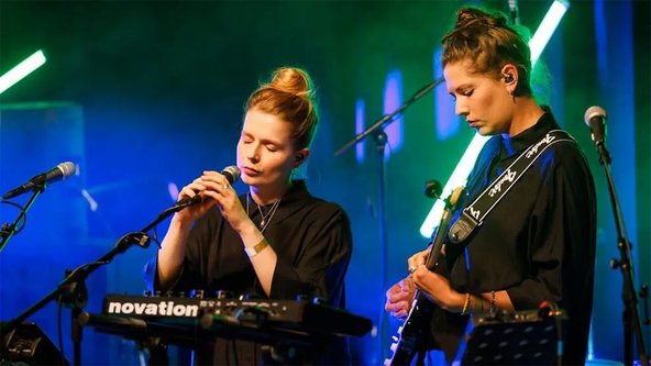 Zwei musizierende Frauen. Eine Frau singt an einem Mikrofon, die andere Frau spielt an einer E-Gitarre.