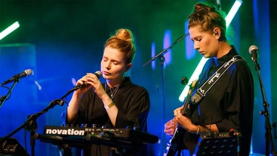 Zwei musizierende Frauen. Eine Frau singt an einem Mikrofon, die andere Frau spielt an einer E-Gitarre.